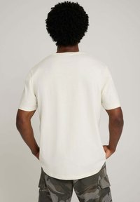 Kremowy T-shirt z krótkim rękawem wykonany z bawełny o luźnym kroju. Posiada okrągły dekolt i lekko zakrzywiony dół.