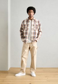 Plaid shirt in beige, blue, and rust; white t-shirt beneath; beige cargo pants; white sneakers; standing against a neutral background.