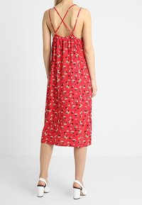 Robe midi rouge à fleurs avec des bretelles spaghetti croisées, un dos froncé et un tissu léger. Présente de petits motifs floraux et une coupe ample.