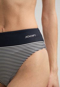 Vrouw in marineblauwe en witte gestreepte hoge taille bikini broekjes met het "JOOP!" logo aan de zijkant tegen een effen achtergrond.