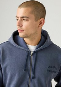 Joven con pelo corto, vestido con una sudadera con cremallera azul marino con cordones y el texto "Two Rodeo Athletic Club", mirando hacia su izquierda ante un fondo neutro.