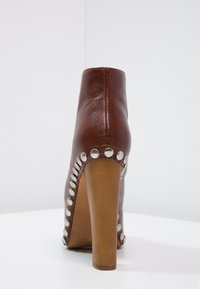 Bota de tobillo de cuero marrón con tacón alto. Presenta tachuelas plateadas a lo largo del costado y un diseño trasero suave y redondeado.