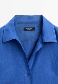 Blaues Leinenhemd mit Kragen, einem einzelnen Knopfverschluss und einer subtilen Textur. Das Markenlabel "Massimo Dutti" ist im Inneren sichtbar.