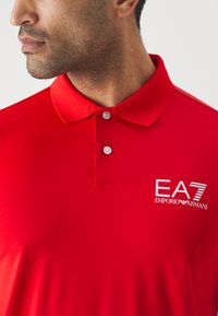 Red polo shirt featuring a buttoned collar, a textured fabric, and a white embroidered logo reading "EA7 Emporio Armani" on the chest.