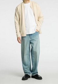 Persona vestida con una chaqueta beige con cremallera sobre una camiseta blanca, jeans de pierna ancha de color azul claro y zapatos negros, de pie con una mano en el bolsillo.