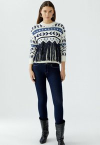 Brede gebreide trui in crème met marineblauwe geometrische patronen en franjes aan de onderkant, gecombineerd met strakke donkerblauwe jeans en zwarte laarzen.