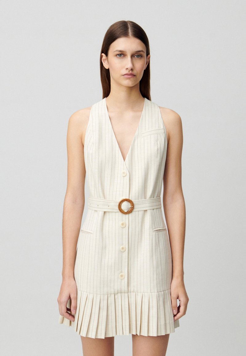 Robe sans manches beige à rayures avec un décolleté en V profond, boutonnée sur le devant et une taille cintrée avec une boucle tissée ronde. Jupette évasée et plissée.