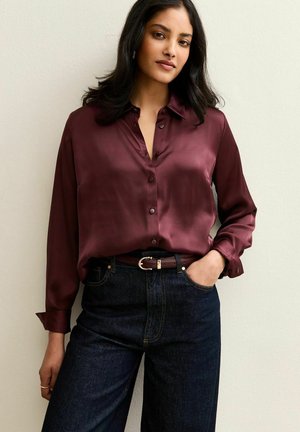 Camisa de satén burdeos con botones, cuello, mangas largas y corte relajado, combinada con unos vaqueros de denim oscuro y un cinturón marrón.