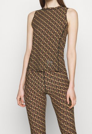 Vrouw met een bruine mouwloze top en bijpassende broek met zwart-rood abstract geometrisch patroon, staand tegen een effen lichte achtergrond.