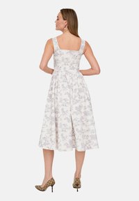 Weißes Midikleid mit floralem Muster, quadratischem Ausschnitt und breiten Trägern. Verfügt über einen fließenden Rock und einen Rückenschlitz. Kombiniert mit gemusterten High Heels.