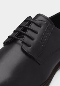 Κοντινή λήψη ενός μαύρου δερμάτινου παπουτσιού με τέσσερις οπές για κορδόνια και μαύρα κορδόνια σε λευκό φόντο.