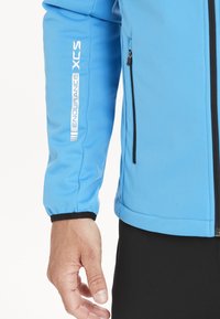 Lyseblå softshell jakke med en lynlåslomme, sorte accenter, justerbare manchetter og trykt "ENDURANCE XCS" branding på ærmet.
