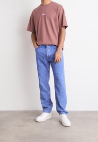 Camiseta de algodón marrón con un pequeño logo rosa en el pecho, combinada con jeans rectos de mezclilla azul claro y zapatillas blancas. Diseño sencillo.