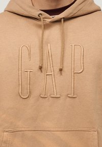 Κοντινό πλάνο ενός μπεζ φούτερ με μεγάλες κεντητές γράμματα "GAP" στο στήθος και κορδόνια που κρέμονται από τη κουκούλα.