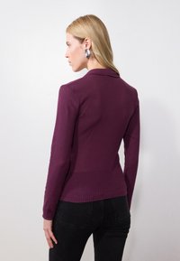 Long-sleeve top in deep purple ribbed fabric, featuring a sharp collar and fitted design, worn with black high-waisted jeans.