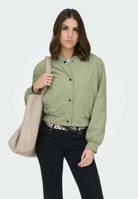 Veste bomber courte vert olive avec fermeture à boutons, poignets élastiques et poches latérales. Associée à un pantalon noir et un sac fourre-tout léger.