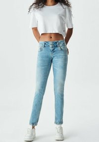 Jean en denim bleu clair à coupe slim, avec une taille haute, une fermeture à deux boutons et un léger délavage. Associé à un t-shirt court en coton blanc.