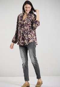 Burgunderfarbene Bluse mit floralem Muster, V-Ausschnitt, langen Ärmeln und lockerer Passform; kombiniert mit grauen Skinny Jeans und beigen Slippern.