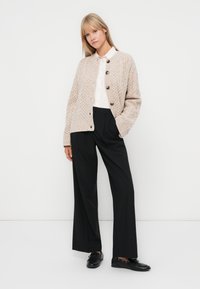 Beige knitted cardigan with textured pattern and five dark buttons, paired with a white collared shirt and black wide-leg trousers.