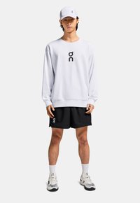 Sweat-shirt blanc avec logo noir, short noir, chaussettes blanches et chaussures de course. La casquette est également blanche avec logo et accents en maille.