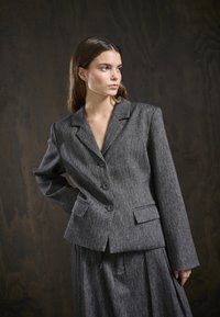 Grau strukturierter Blazer mit Reverskragen, zwei Knopfverschluss und frontalen Taschen. Kombiniert mit einem passenden grauen Rock.