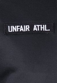 Pezzo di tessuto nero con la scritta "UNFAIR ATHL." in bold lettere bianche. Lo sfondo ha una finitura texture.