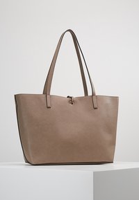 Ruskea keinonahka-tote laukku, jossa on kaksi pitkää kantohihnaa, yksinkertainen suorakulmainen muoto ja minimalistische muotoilu, jossa on keskellä oleva korostus.