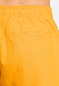 Gelbe Badeshorts aus leichtem Material, mit elastischem Taillenbund und einer kleinen Gesäßtasche. Glatte Textur, einfaches Design.