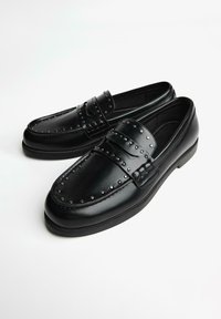 Fekete bőr mokaszinok díszítő szegecsekkel a felsőrész mentén, sima felülettel és gumitalppal. Klasszikus dizájn dupla pánt díszítéssel.