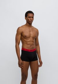 Μαύρα ανδρικά εσώρουχα boxer με κόκκινη ελαστική μέση που φέρει το λογότυπο "HUGO". Λείο ύφασμα, εφαρμοστό σχέδιο, κομμένα πάνω από τη μέση του μηρού.