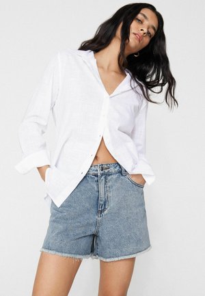 Frau mit langen dunklen Haaren, die ein lockeres weißes Hemd mit Knöpfen und hoch taillierte blaue Jeansshorts mit ausgefransten Säumen trägt, Hände in den Taschen.