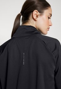 Giacca sportiva nera con colletto alto e schiena texturizzata, caratterizzata da una linea verticale di dettagli riflettenti. Tessuto liscio e leggero.