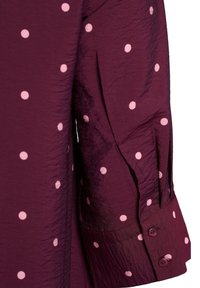 Nahaufnahme eines strukturierten dunkelvioletten Stoffes mit hellrosa Polka-Dots und einem Ärmelbündchen mit zwei Knöpfen.