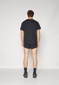 Camiseta negra de manga corta combinada con pantalones cortos negros que presentan líneas de acento amarillas. El modelo lleva calcetines negros y zapatillas.