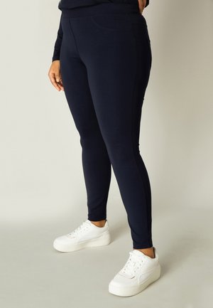 Marineblauwe stretchleggings met een nauwsluitend ontwerp, voorzien van een tailleband, zijzakken en een gladde textuur, gecombineerd met witte sneakers.