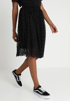 Personne portant une jupe midi noire à pois et des baskets noires et blanches, marchant avec un pied légèrement levé.