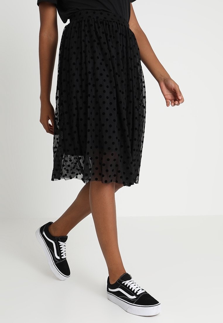 Personne portant une jupe midi noire à pois et des baskets noires et blanches, marchant avec un pied légèrement levé.