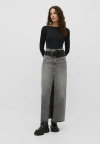 Schwarzes Langarmoberteil kombiniert mit einem grauen Denim-Maxirock, der einen V-Ausschnitt hat und mit einem breiten schwarzen Gürtel und einer großen silbernen Schnalle gesichert ist.