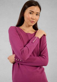 Giovane donna che indossa un maglione viola a maniche lunghe con bottoni di perla ai polsini, in piedi con le braccia incrociate contro uno sfondo grigio uniforme.