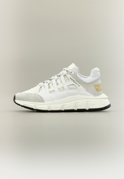 Baskets Versace blanches et grises avec un design à lacets, semelle épaisse ornée d'un motif clé grec, et détail du logo doré sur le talon.