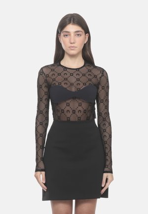 Vrouw met lang donker haar die een zwarte doorschijnende top met lange mouwen en patroon draagt over een zwarte bandeau en een zwarte rok met hoge taille.