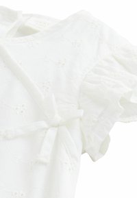Haut en coton blanc avec des manches courtes à volants, broderie florale, et un détail à nouer au niveau du col. Texture douce, design léger.