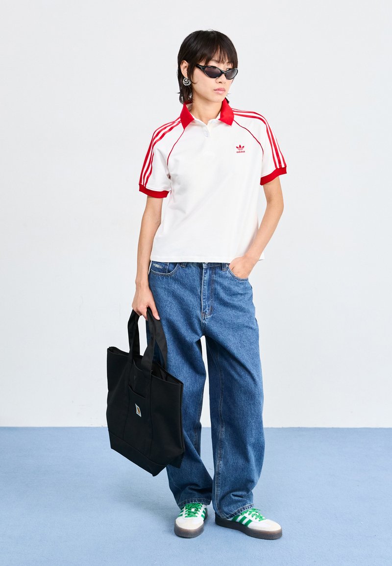 Biała koszulka polo z czerwonymi akcentami i wzorem w trzy paski, zestawiona z luźnymi niebieskimi dżinsami i zielonymi trampkami w paski. Niesie czarną torbę na ramię.