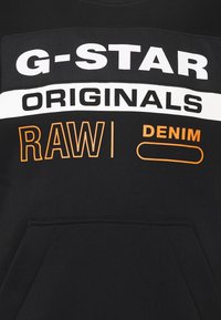 Tissu noir avec texte blanc et orange "G-STAR ORIGINALS RAW | DENIM" au-dessus d'une poche kangourou.