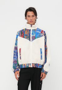 Fleece jacket with a high collar, featuring a white body and colorful patterned sleeves. Zippered front and side pockets.