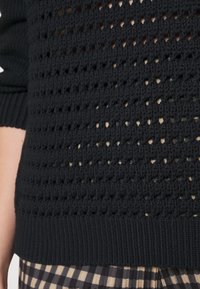 Svart stickad tröja med en lös, öppen struktur och ribbat avslut. Materialet verkar texturerat och visar en subtilt lager-på-lager-design.