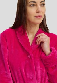 Bata de felpa fucsia con cuello acolchado, botones y puños anchos. Textura suave y cinturón en la cintura para un ajuste seguro. Detalles visibles en el cuello y los puños.