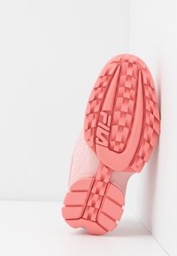 Semelle d'une basket Fila rose avec motif de bande de roulement texturé, inclinée verticalement contre un mur d'angle blanc et un sol.