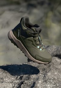 Căciulă de drumeție verde, cu un strat superior texturat, secțiuni întărite, design ușor și o talpă durabilă. Sunt utilizate materiale din suede și sintetic.