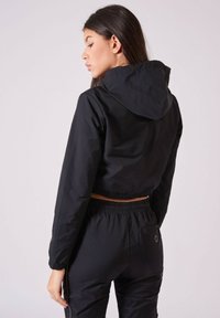 Schwarze, kurze Jacke mit Kapuze, aus glattem Material. Mit elastischen Bündchen und einem verstellbaren Saum, kombiniert mit passenden schwarzen Hosen.